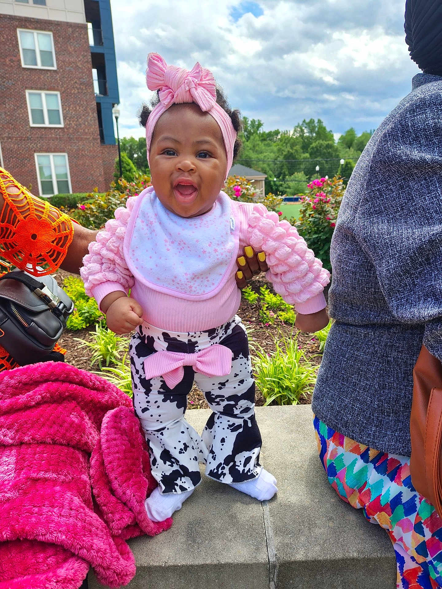 Shawty-Zuri joined the competition — help win amazing prizes! baby, toddler, child, outdoor, smiling, pink_headband, pink_top, cow_print_pants, bow, concrete, hand_support, plants, flowers, blanket, adult_hand, cloudy_sky, building, grass, person, colorful_clothing