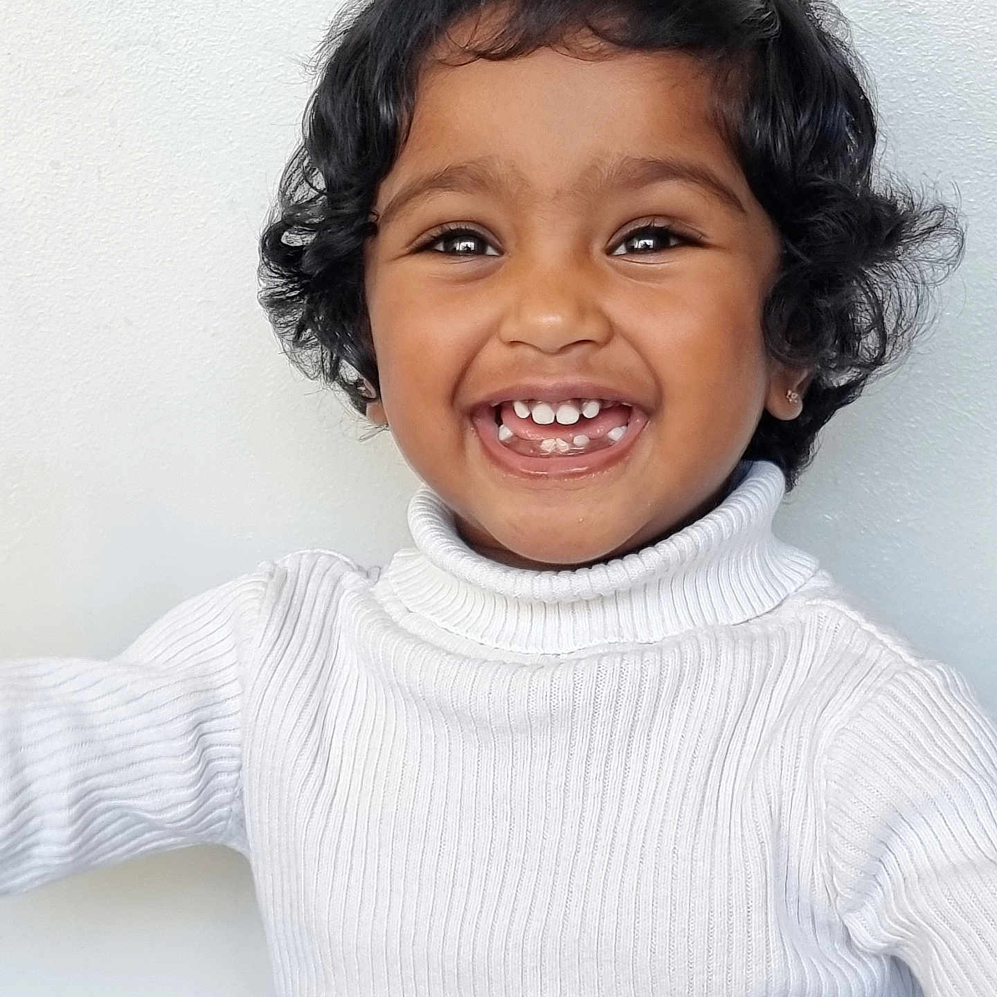 Aathira is registered to the contest to win money with this photo: apparel, child, closeup, curly_hair, cute, face, fashion, happy, indoor, innocence, joyful, light_background, person, portrait, smiling, teeth, toddler, turtleneck, white_sweater, young_child
