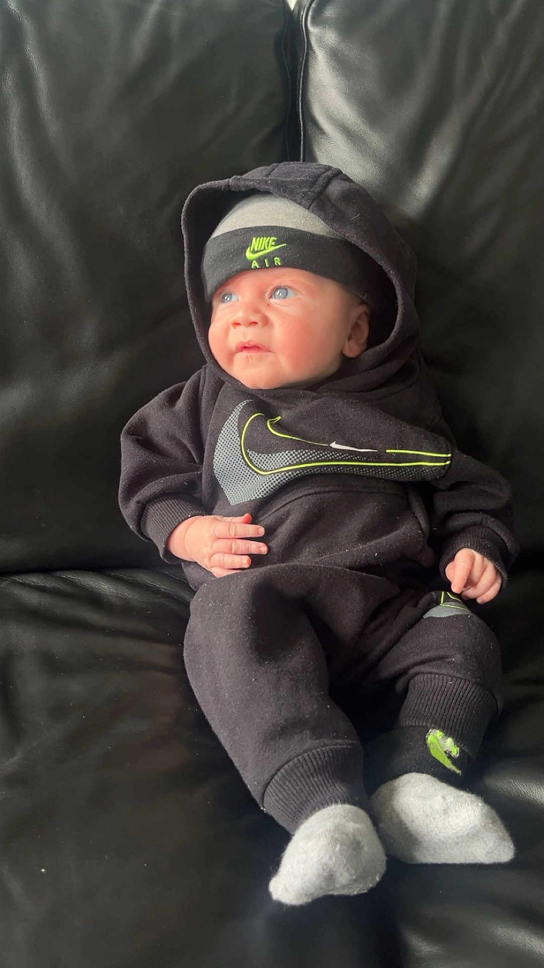 Roman is registered to the contest to win money with this photo: baby, infant, black_clothing, nike, hoodie, hat, socks, couch, leather, blue_eyes, cute, portrait, indoor, child, small_hand, sitting, looking_away, cozy, casual, comfortable