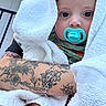 Lane Frost Chapman is registered to the contest to win money with this photo: arm, baby, blanket, child, close_up, cute, expression, face, hand, holding, indoor, infant, pacifier, person, portrait, skin, tattoo, towel, wrapped, young