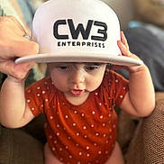 Lane Frost Chapman joined the competition — help win amazing prizes! baby, blurred_background, cap, child, couch, curious, cute, face, hat, holding_hat, indoor, infant, onesie, person, portrait, red_clothing, sitting, small_hands, white_text, young_child