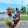 beard, car, casual_clothing, daylight, dog, goatee, greenery, holding, man, outdoor, parking_lot, puppy, sidewalk, sky, smiling, stop_sign, street_lamp, tattoo, tie_dye_shirt, trees
