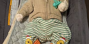 Weston is registered to the contest to win money with this photo: baby, infant, hoodie, striped_pants, cartoon_bear, slippers, pacifier, baby_seat, quilted_pad, cozy, cute, child, indoor, clothing, footwear, person, face, lying_down, warm, soft