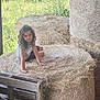 child, girl, hay_bale, hay, barn, wooden_pallet, curly_hair, smiling, outdoor, rural, field, greenery, nature, sunlight, sitting, casual_clothing, playful, farm, summer, relaxed