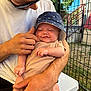 baby, man, hat, outdoor, smile, sunglasses, hand, fence, table, grass, shirt, person, holding, infant, skin, arm, face, daylight, expression, casual