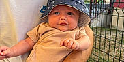 Tymëlio participe au concours pour gagner de l'argent avec cette photo : baby, child, infant, bucket_hat, onesie, smiling, person, adult, arm, hand, outdoor, fence, grass, table, tshirt, nike, sunlight, face, seated, building