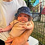 baby, child, infant, bucket_hat, onesie, smiling, person, adult, arm, hand, outdoor, fence, grass, table, tshirt, nike, sunlight, face, seated, building