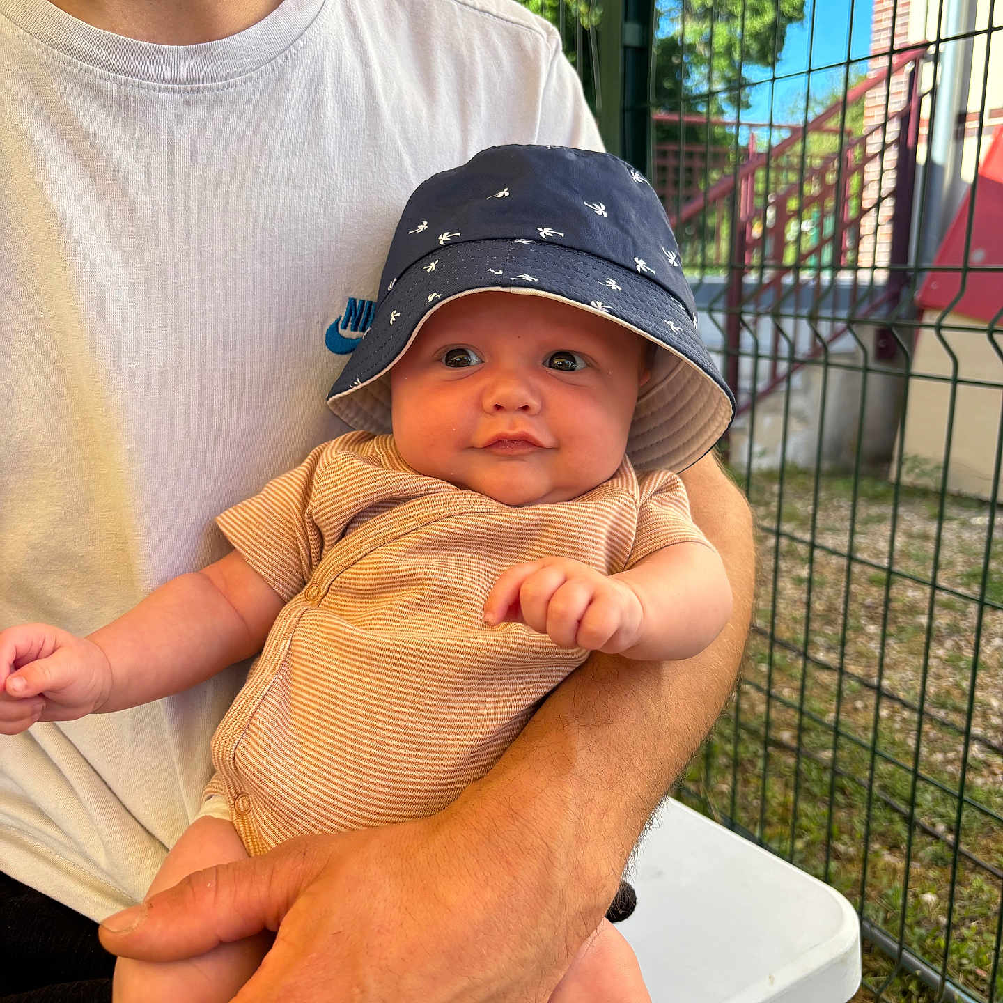 Tymëlio participe au concours pour gagner de l'argent avec cette photo : adult, arm, baby, bucket_hat, building, child, face, fence, grass, hand, infant, nike, onesie, outdoor, person, seated, smiling, sunlight, table, tshirt