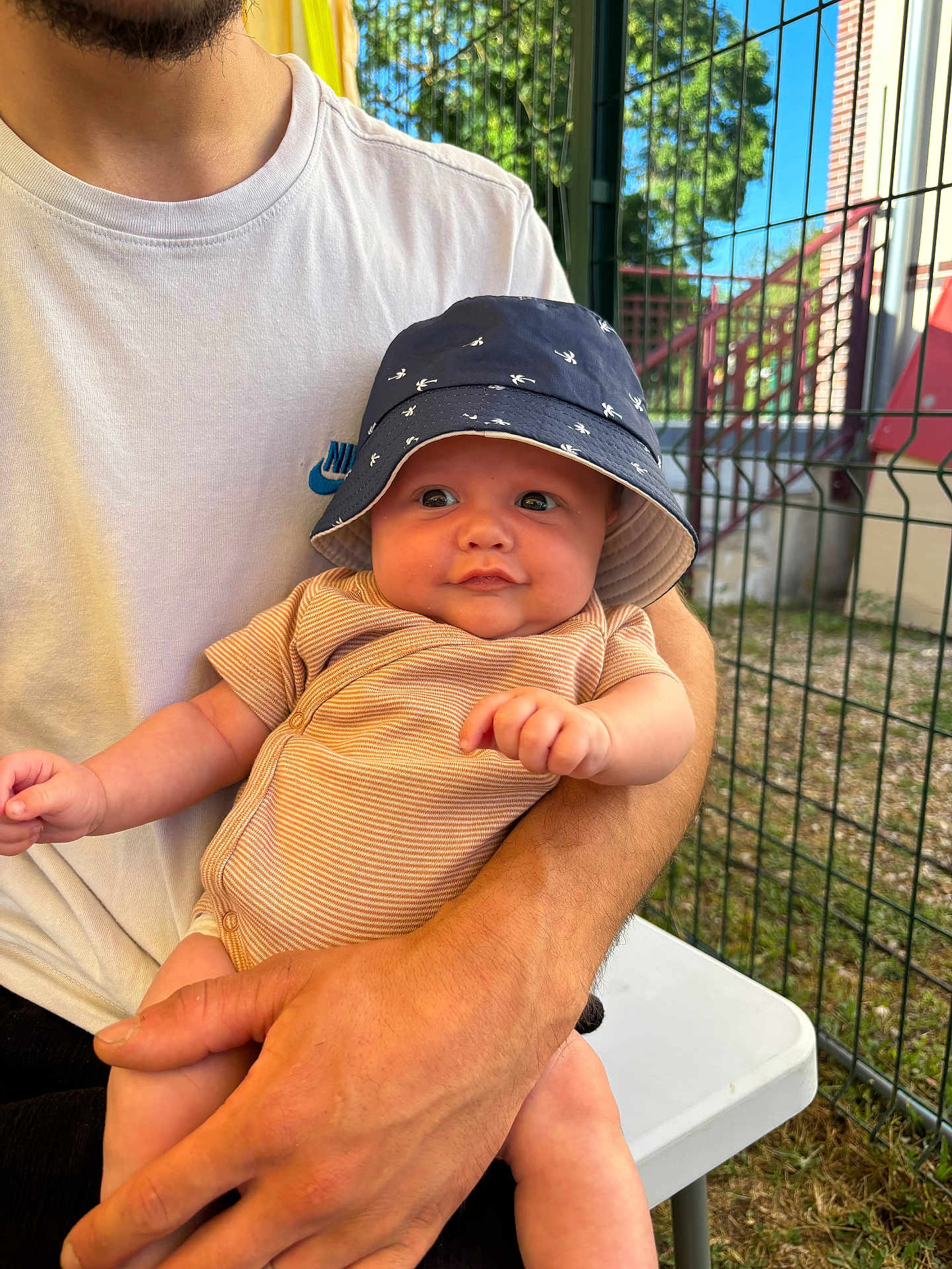 Tymëlio participe au concours pour gagner de l'argent avec cette photo : baby, child, infant, bucket_hat, onesie, smiling, person, adult, arm, hand, outdoor, fence, grass, table, tshirt, nike, sunlight, face, seated, building