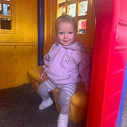 Alaura-Jane is registered to the contest to win money with this photo: toddler, child, playhouse, plastic, purple_hoodie, striped_pants, white_shoes, indoor_play_area, smiling, seated, colorful, yellow_wall, red_frame, window, playground, young_child, casual_clothing, happy, cute, small