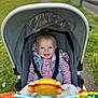 toddler, child, stroller, smile, blue_eyes, outdoor, pathway, grass, toy, colorful, coat, jacket, blanket, safety_strap, baby_toy, happy, portrait, person, daylight, casual