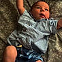 baby, infant, child, bed, pillow, blue_shirt, shorts, floral_pattern, raised_arm, barefoot, lying_down, indoor, curious_expression, fabric, bedspread, soft_light, skin, human, person, casual_clothing