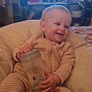 Lyla is registered to the contest to win money with this photo: baby, child, bottle, pajamas, smile, indoor, television, cushion, furniture, screen, happy, person, infant, clothing, living_room, relaxing, seat, home, technology, background