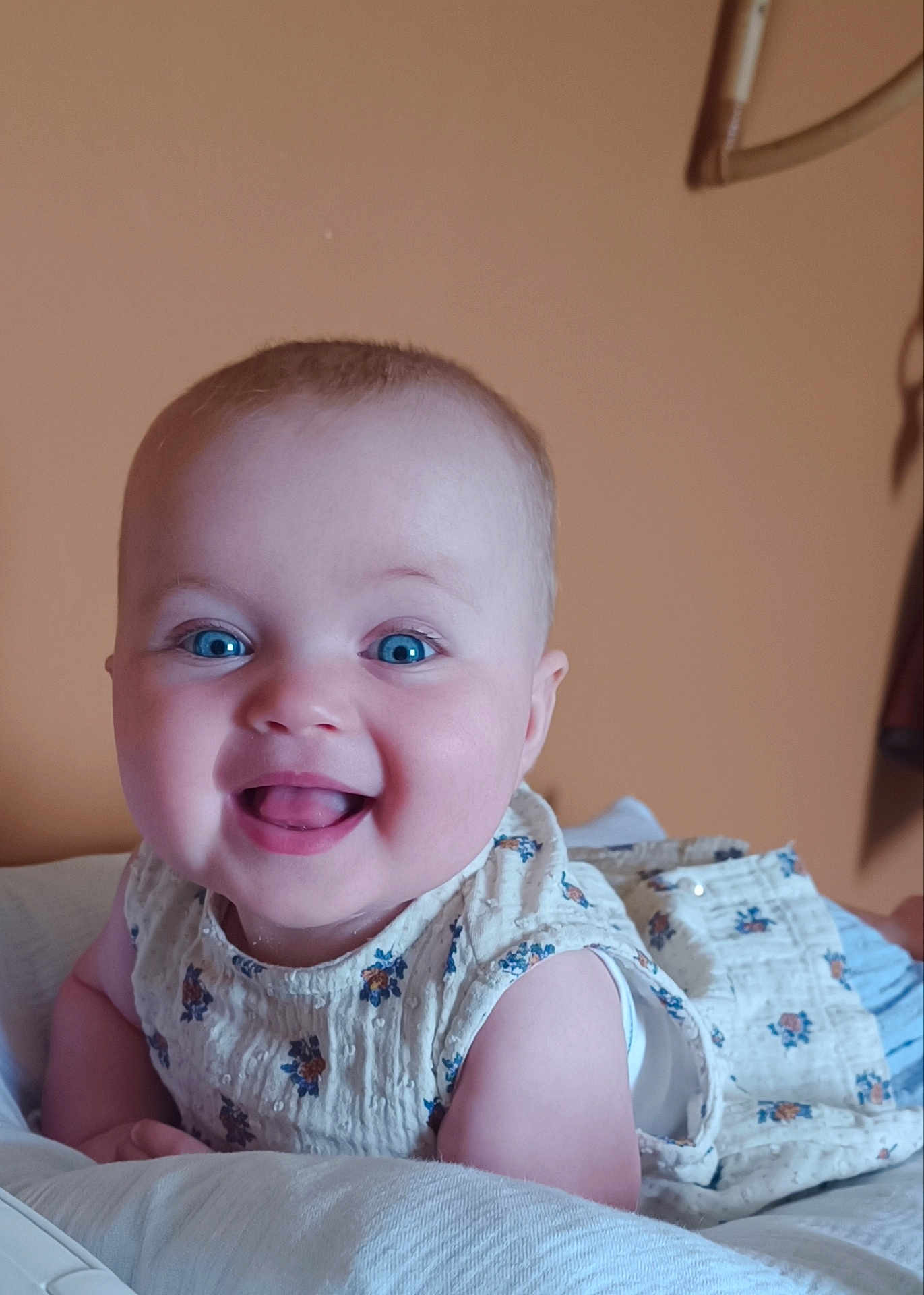 Margot participe au concours pour gagner de l'argent avec cette photo : baby, smiling, blue_eyes, floral_dress, infant, happy, cute, child, portrait, indoors, soft_lighting, skin, face, person, toddler, lying_down, white_surface, warm_background, cheerful, closeup