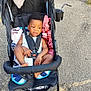 baby_clothes, blanket, casual, child, cup_holder, curly_hair, expression, footwear_free, grass, outdoor, pavement, plastic_cup, seatbelt, sidewalk, stroller, summer, sunlight, toddler, towel, wheels