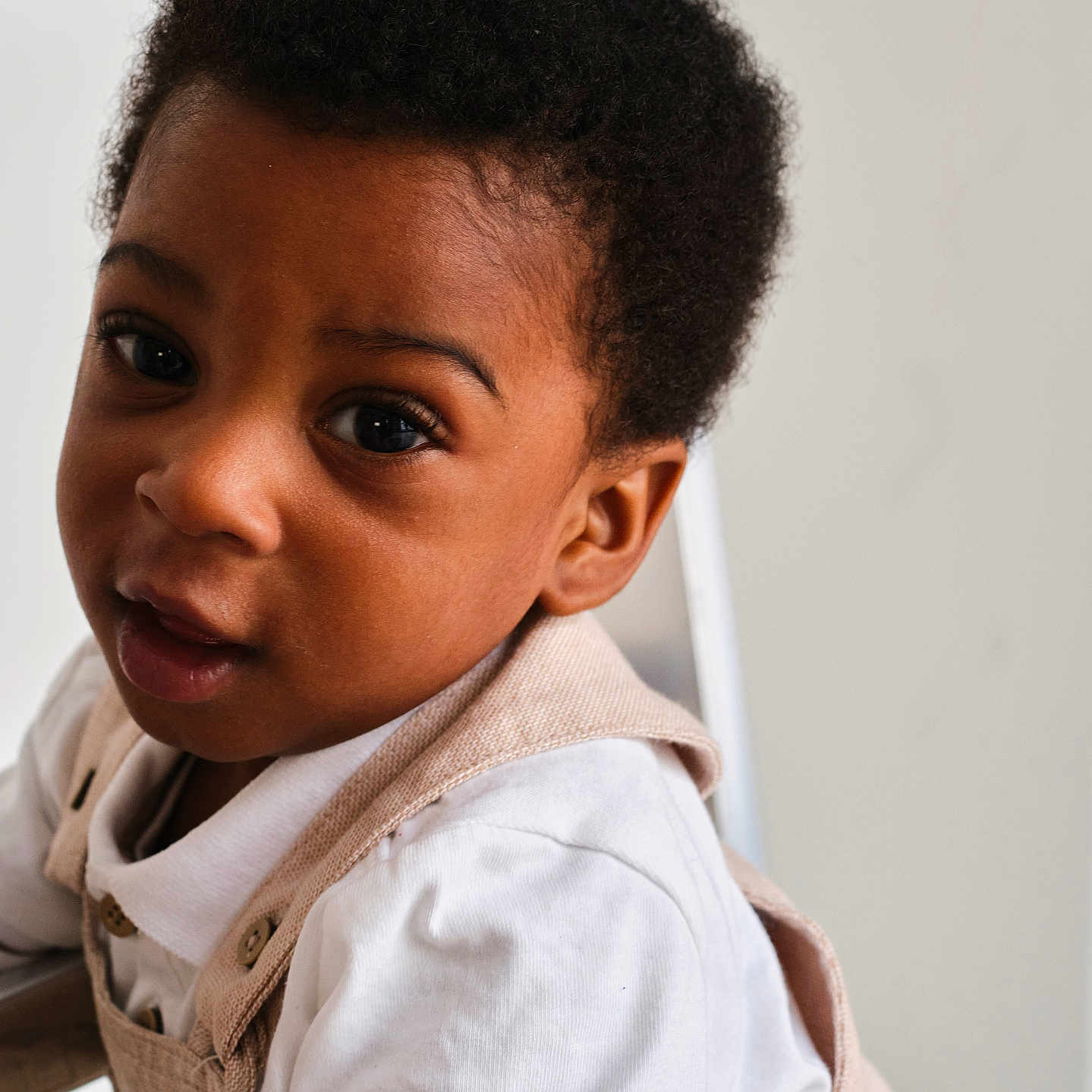 Kris-Emmanuel participe au concours pour gagner de l'argent avec cette photo : child, close_up, clothing, curious, curly_hair, cute, expression, eyes, face, gentle, head, indoor, neutral_background, overalls, person, portrait, skin, toddler, white_shirt, young_child