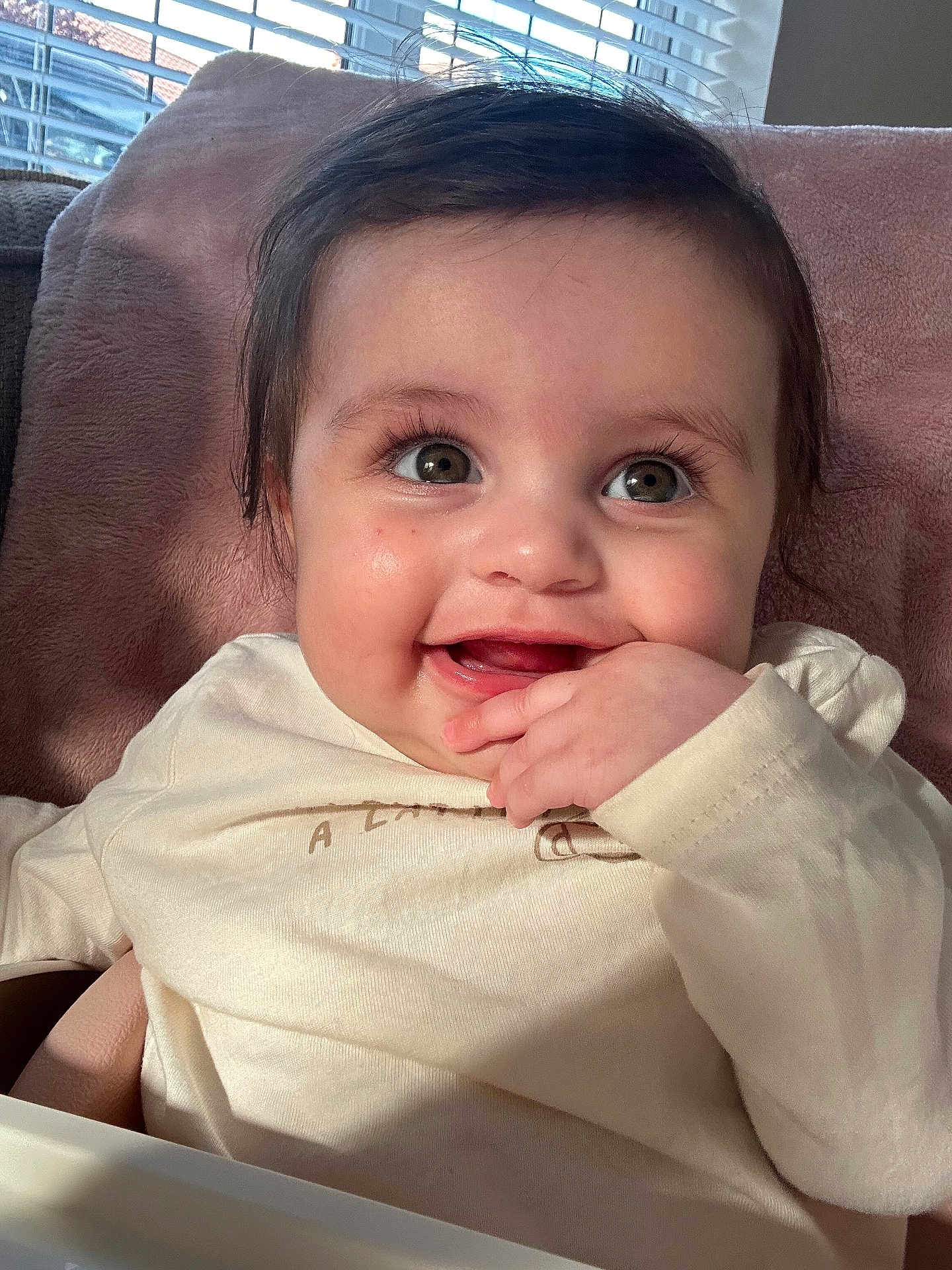 Mila is registered to the contest to win money with this photo: baby, child, smile, face, hand, eyes, cushion, soft_fabric, indoor, window, sunlight, shadow, long_eyelashes, clothing, cheeks, portrait, happy, infant, cute, close_up