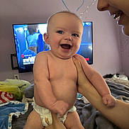 Grayson is registered to the contest to win money with this photo: baby, diaper, smile, happy, person, face, hands, adult, bed, blanket, television, room, indoor, joyful, candid, holding, home, expression, child, parent