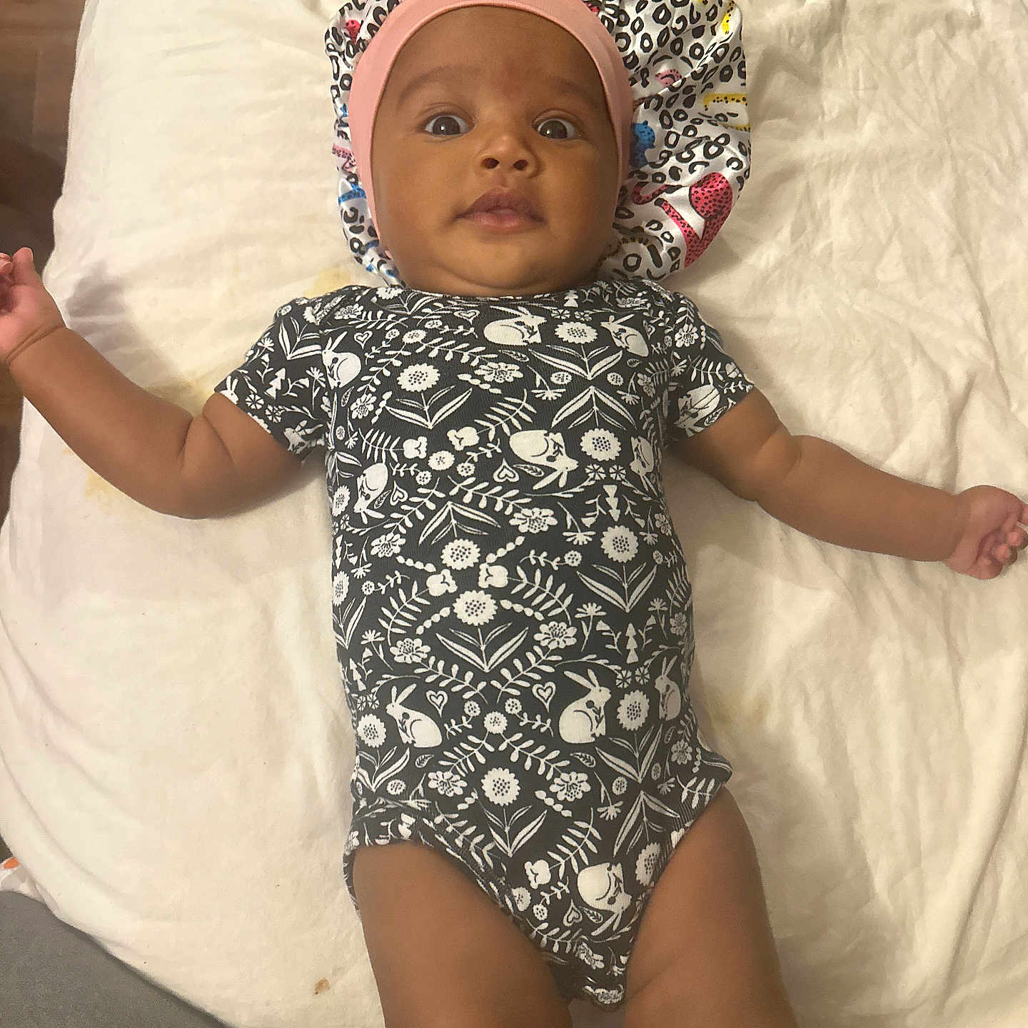 Essence joined the competition — help win amazing prizes! baby, infant, onesie, headband, lying_down, curious, indoor, blanket, soft_surface, patterned_clothing, arms_outstretched, child, person, cute, young_child, baby_clothes, cozy, resting, looking_at_camera, home