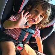 Ryleigh is registered to the contest to win money with this photo: child, toddler, car_seat, safety_harness, seatbelt, car_interior, sunlight, curly_hair, pink_checkered_shirt, black_shorts, hand, face, blue_eyes, smiling, cute, person, young_child, seat, window, daylight