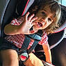 child, toddler, car_seat, safety_harness, seatbelt, car_interior, sunlight, curly_hair, pink_checkered_shirt, black_shorts, hand, face, blue_eyes, smiling, cute, person, young_child, seat, window, daylight