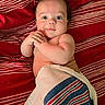 baby, infant, towel, striped_blanket, red_background, lying_down, cute, chubby_cheeks, hands, face, portrait, skin, person, child, indoors, expression, eyes, smile, newborn, resting