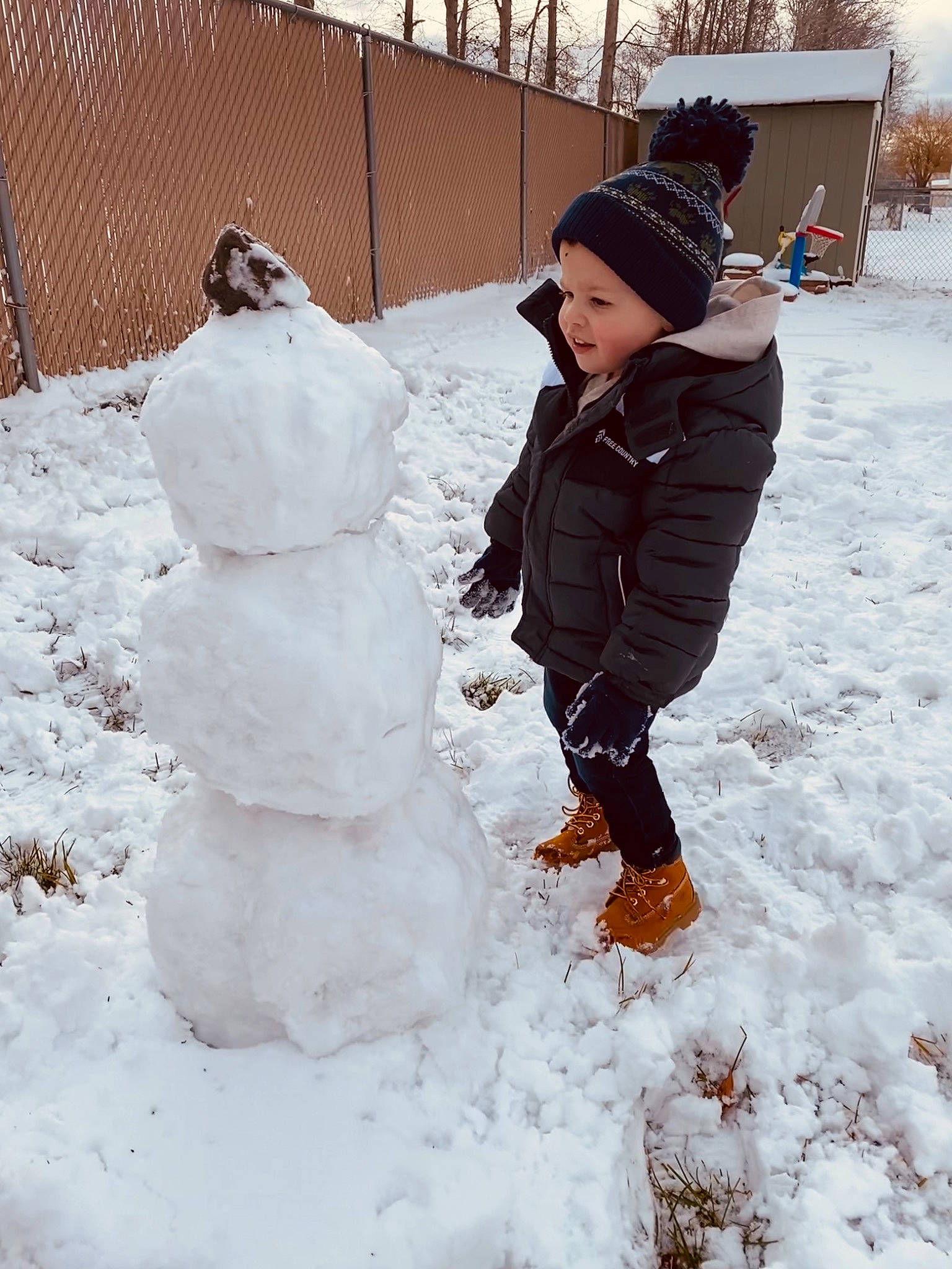 Flynn is registered to the contest to win money with this photo: baby_toddler_clothing, child, face, fence, freezing, fun, fur, hat, headwear, parka, people_in_nature, person, playing_in_the_snow, playing_with_kids, precipitation, snow, snow_boot, snowman, toddler, winter