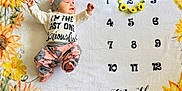 Gemma is registered to the contest to win money with this photo: baby, infant, milestone_blanket, sunflower, flower_crown, months, hat, clothing, lying_down, cute, pink_pants, white_shirt, numbers, yellow_flowers, patterned_pants, baby_milestone, portrait, child, soft_blanket, expression