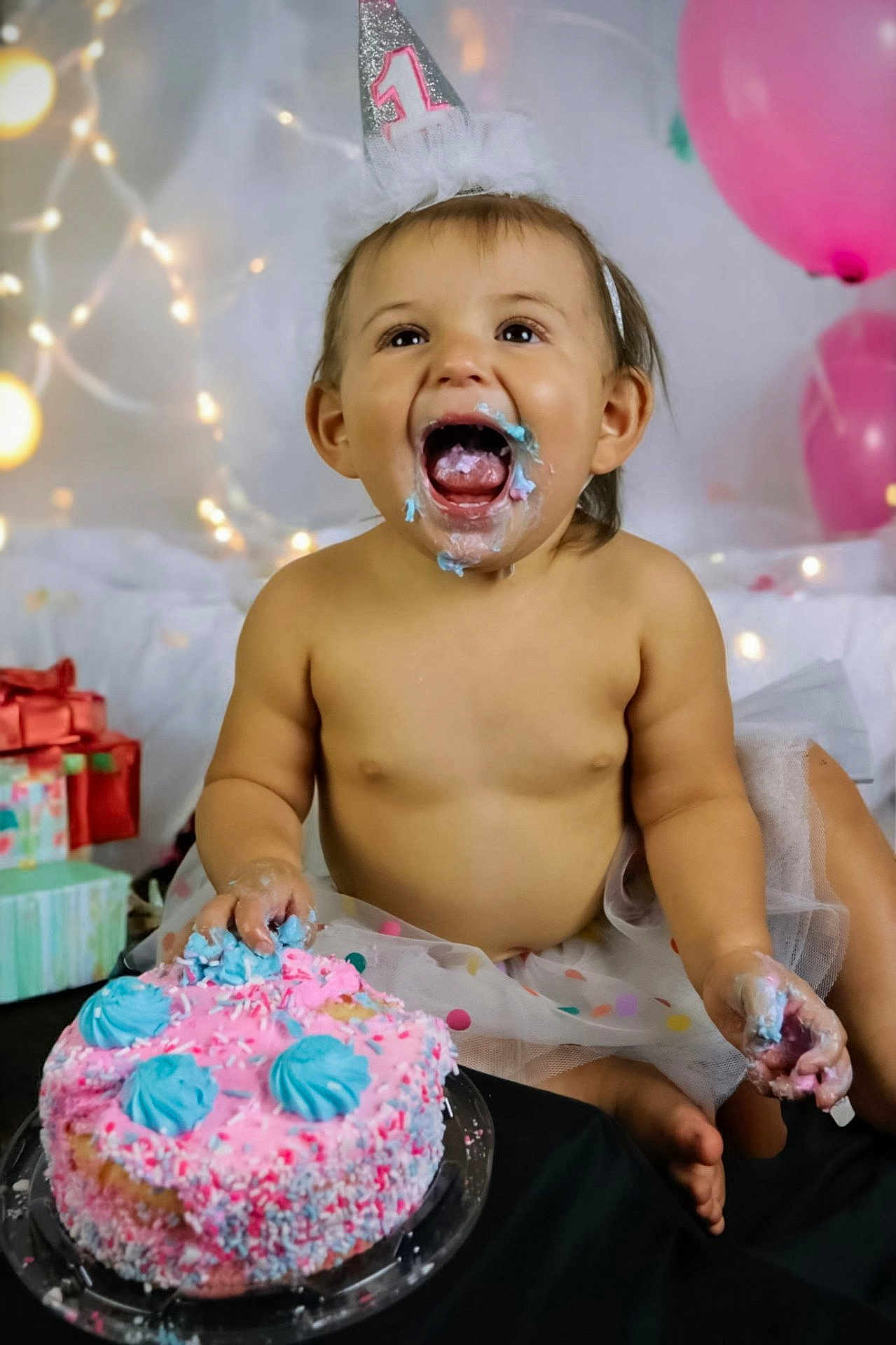Rileigh is registered to the contest to win money with this photo: baby, birthday_hat, cake, pink_cake, blue_frosting, smiling, face_paint, party, celebration, balloons, presents, tutu, messy, happy, child, festive, sitting, first_birthday, indoor, colorful