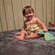 Rileigh is registered to the contest to win money with this photo: toddler, child, smiling, sitting, toy, pop_it, blanket, romper, yellow_clothing, indoor, wood_panel_wall, happy, barefoot, playful, person, infant, cute, young_child, home_interior, portrait