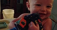 Carson is registered to the contest to win money with this photo: baby, child, smiling, indoor, portrait, high_chair, stuffed_toy, blanket, blue_blanket, home, living_room, furniture, window, curtains, cabinet, bucket, hand, sitting, cozy, family_photo
