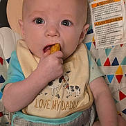 Carson is registered to the contest to win money with this photo: baby, infant, child, portrait, sitting, bouncer, bib, snack, eating, hand, face, eyes, smiling, indoor, close_up, colorful, patterned, baby_gear, safety_label, adorable