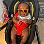 baby, child, sunglasses, red_onesie, car_seat, indoor, floor, mop, dustpan, happy, smiling, cute, infant, legs, feet, hands, bib, black_car_seat, wood_floor, playful