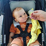 Scott joined the competition — help win amazing prizes! baby, black_stroller, blanket, child, childcare, curious_expression, cute, daylight, feeding, feeding_time, hand, infant, orange_clothing, outdoor, portrait, seatbelt, sitting, spoon, stroller, yellow_blanket
