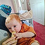 baby, infant, banana, hand, orange_clothing, couch, living_room, door, blanket, carpet, person, furniture, indoor, feeding, cute, child, face, skin, nail_polish, ring