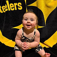 Ellie joined the competition — help win amazing prizes! baby, child, smiling, sitting, clothing, blanket, football_team_logo, black, yellow, happy, cute, portrait, indoors, feet, hands, face, expression, person, toddler, hair