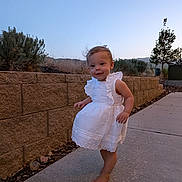 Blake is registered to the contest to win money with this photo: barefoot, bush, child, cute, evening, grass, happy, nature, outdoor, person, portrait, sidewalk, sky, smiling, stone_wall, toddler, tree, walking, white_dress, young_child