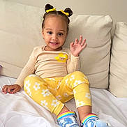 Alana joined the competition — help win amazing prizes! child, toddler, smiling, waving, yellow_clothing, floral_pattern, hair_buns, hair_bows, blue_crocs, rainbow_shoes, sitting, couch, white_bedding, indoor, happy, cute, young_child, home, casual_clothing, portrait