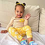 child, toddler, smiling, waving, yellow_clothing, floral_pattern, hair_buns, hair_bows, blue_crocs, rainbow_shoes, sitting, couch, white_bedding, indoor, happy, cute, young_child, home, casual_clothing, portrait
