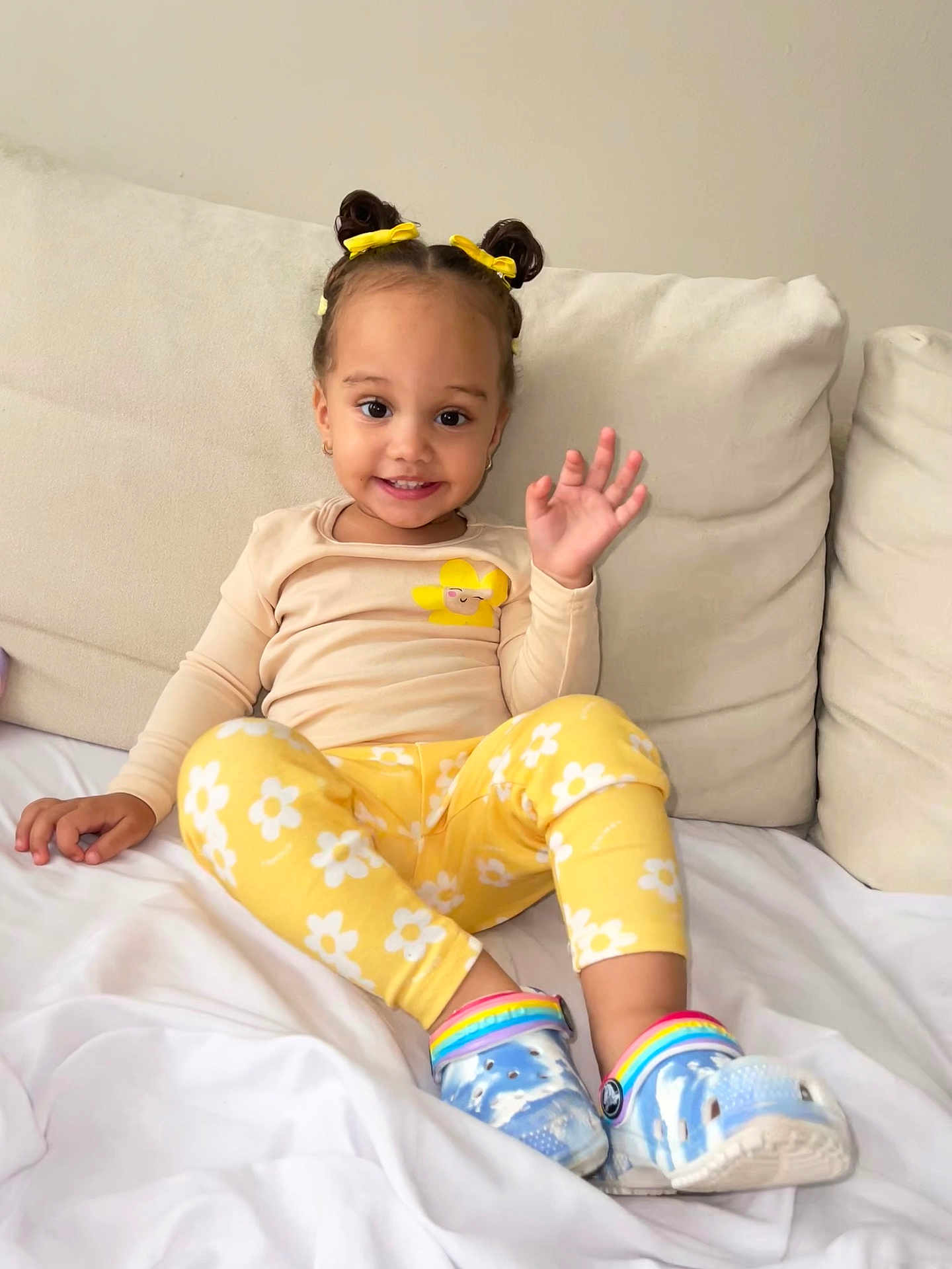 Alana joined the competition — help win amazing prizes! child, toddler, smiling, waving, yellow_clothing, floral_pattern, hair_buns, hair_bows, blue_crocs, rainbow_shoes, sitting, couch, white_bedding, indoor, happy, cute, young_child, home, casual_clothing, portrait