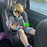 Jaxon is registered to the contest to win money with this photo: child, car_seat, sunglasses, toy, car_interior, seatbelt, casual_clothing, shorts, tshirt, red_shoes, reflection, window, daylight, road, pout, blond_hair, sitting, playful, child_safety, vehicle