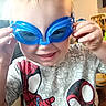 child, goggles, blue_goggles, smiling, indoor, spiderman_shirt, tshirt, hands, blond_hair, face, person, playful, furniture, chair, basketball, home, young_child, cute, happy, portrait