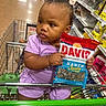 toddler, child, shopping_cart, grocery_store, bag, sunflower_seeds, purple_clothing, hair_bows, sitting, aisle, floor, shelves, products, footwear, curious, holding, portrait, indoor, person, shopping
