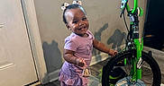 Sahara Jean joined the competition — help win amazing prizes! toddler, child, smiling, purple_dress, crocs, indoor, bicycle, green_bike, wood_floor, door, window, toy, bell, happy, person, flooring, hair_bows, shadow, wall, play