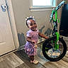 toddler, child, smiling, purple_dress, crocs, indoor, bicycle, green_bike, wood_floor, door, window, toy, bell, happy, person, flooring, hair_bows, shadow, wall, play