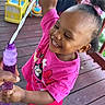 child, toddler, bubble_wand, pink_clothing, happy, smiling, outdoor, deck, toy, play, sandals, person, hair_bow, curly_hair, summer, fun, family, hand, wood_floor, colorful