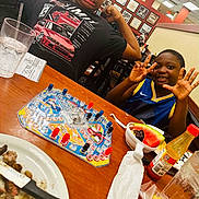 Samir is registered to the contest to win money with this photo: board_game, casual, child, flexing, food, funny_face, glass, happy, hot_sauce, indoor, kids, knife, napkin, person, plate, playing, restaurant, smiling, table, watermelon