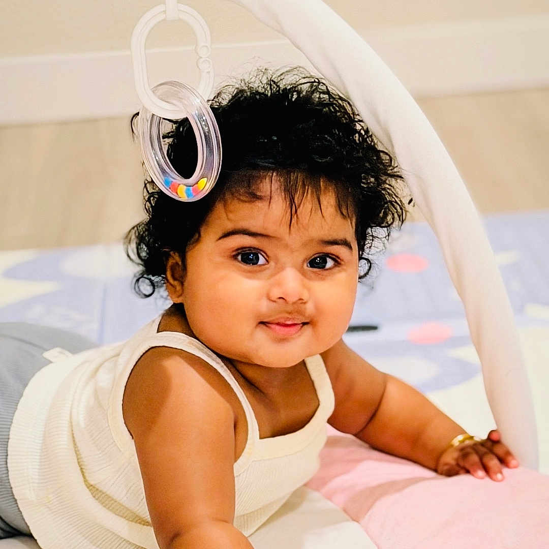Anika Yenkarla is registered to the contest to win money with this photo: arm, baby, bracelets, child, closeup, curly_hair, cute, floor, hand, happy, indoor, infant, lying_down, play_mat, portrait, skin, smiling, soft_toy, toy, white_top