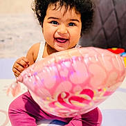 Anika is registered to the contest to win money with this photo: baby, balloon, barefoot, child, clothing, curly_hair, cute, happy, heart_pattern, home, indoors, infant, person, pink, play_mat, purple_pants, sitting, smiling, toy, white_top