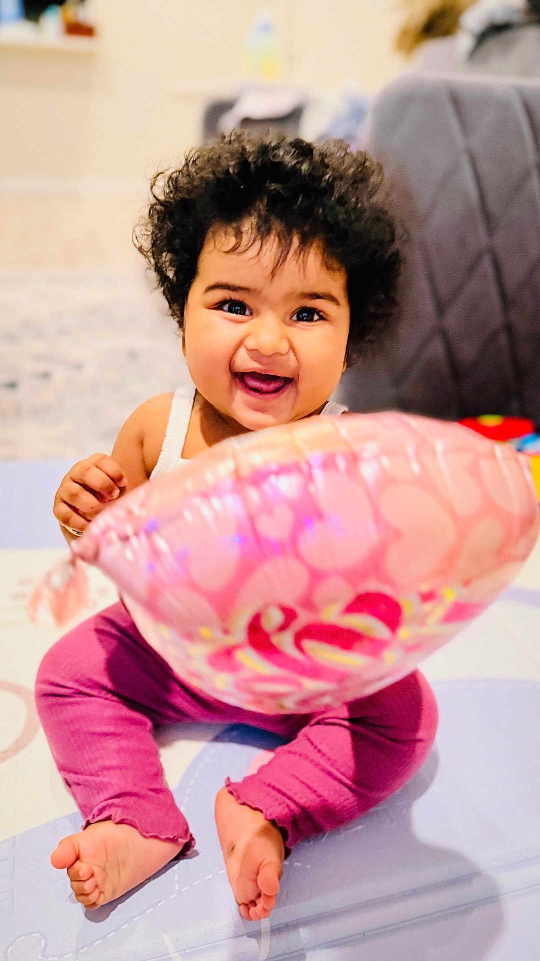 Anika is registered to the contest to win money with this photo: baby, balloon, barefoot, child, clothing, curly_hair, cute, happy, heart_pattern, home, indoors, infant, person, pink, play_mat, purple_pants, sitting, smiling, toy, white_top